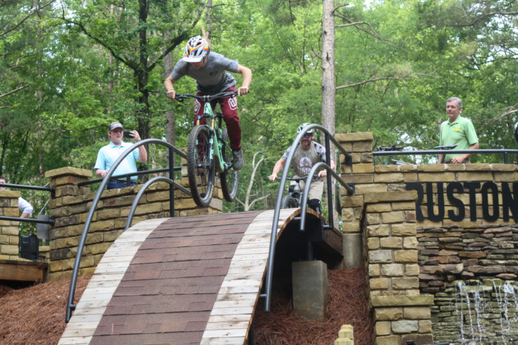 A mountain biker performing a jump over a wooden ramp with curved railings at a bike park, while spectators watch in the background amidst a lush green setting.