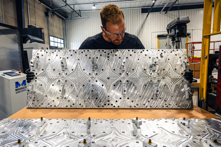 A person wearing safety goggles and gloves inspects a large, intricately machined aluminum plate featuring a decorative pattern, while standing in a workshop filled with industrial machinery. In the foreground, a second, similar aluminum piece is placed on a workbench, which also shows machining fixtures. The environment is well-lit, showcasing a clean and organized manufacturing setting.