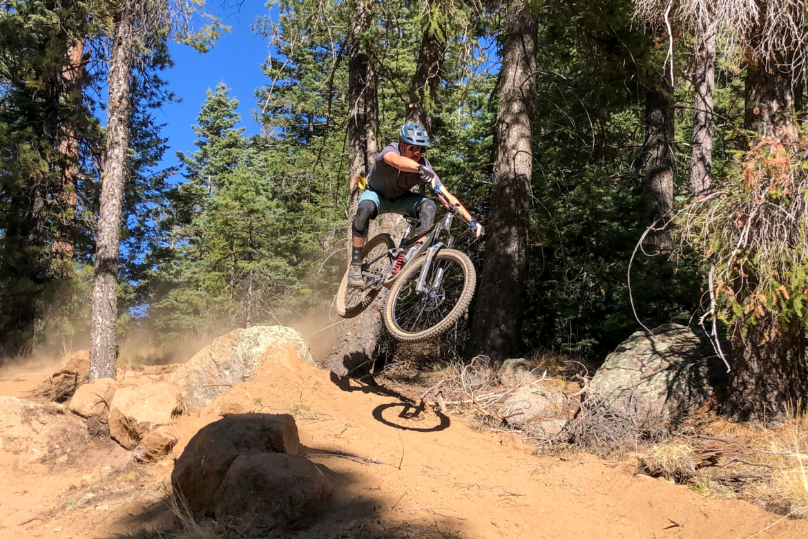 A mountain biker performs a jump over a rocky section of a dirt trail surrounded by tall trees and blue sky, creating a cloud of dust. Full Sail mountain bike trail.