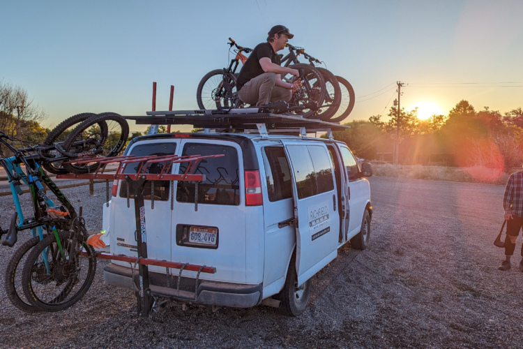 A white van parked on a gravel surface with several mountain bikes secured on a roof rack and a bike rack attached to the back. A person stands on the roof, adjusting the bikes, while another person in a plaid shirt stands nearby, holding a helmet. The sun is setting in the background, casting warm light on the scene. Trees are visible in the distance, creating a natural backdrop.