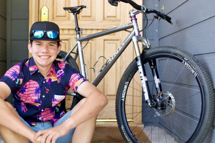 A smiling young person sits on the steps in front of a wooden door, wearing a colorful patterned shirt and baseball cap. Next to them is a silver mountain bike, showcasing its front wheel and handlebars. The background features dark gray wooden siding.
