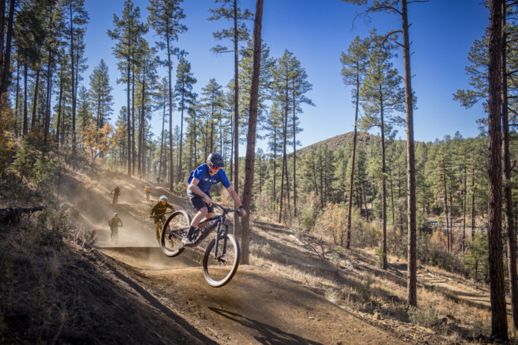 A mountain biker performs a jump on a dirt trail surrounded by tall pine trees, with dust clouds rising from the ground. In the background, other cyclists are riding along the trail, set against a clear blue sky and a rugged mountain landscape.