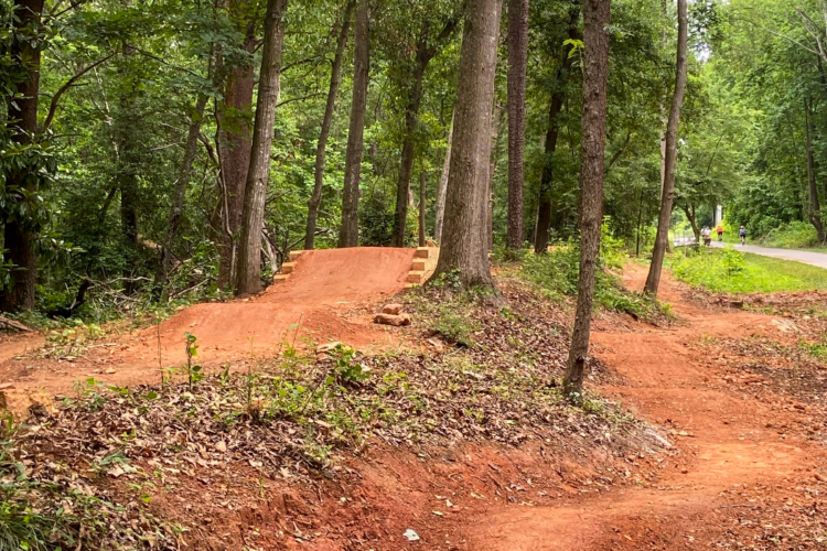A dirt bike trail in a wooded area, featuring a small jump made of compacted earth and wooden ramps. Surrounding the trail are trees, shrubs, and a clear pathway in the distance where two people are walking. The ground is covered with leaves and dirt, contributing to a natural outdoor setting.