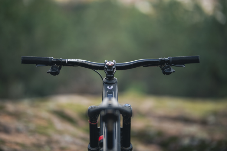Close-up view of a mountain bike's handlebars and front fork, set against a blurred natural background. The handlebars are black, featuring grips and controls, with the bike frame visible extending toward the camera.