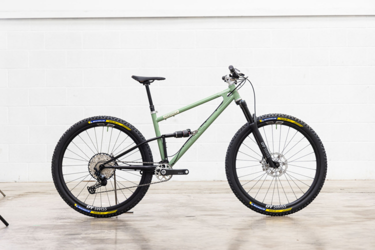 A modern mountain bike with a green frame and black components, showcased against a white wall. The bike features thick tires, a dropper seat post, and a visible drivetrain system. The wheels are outfitted with DT Swiss rims and Michelin tires, indicating performance suitability for off-road trails.