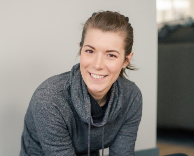 Smiling woman wearing a gray hoodie, sitting on a floor near a light-colored wall. She has her hair pulled back and is looking directly at the camera with a friendly expression. A blurred background indicates a cozy indoor setting.