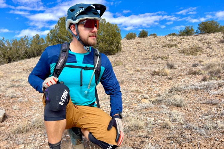 A person wearing a helmet and sunglasses is kneeling on rocky terrain, dressed in a blue long-sleeve shirt and brown shorts, with protective knee pads. They are surrounded by sparse vegetation and a clear blue sky with scattered clouds.