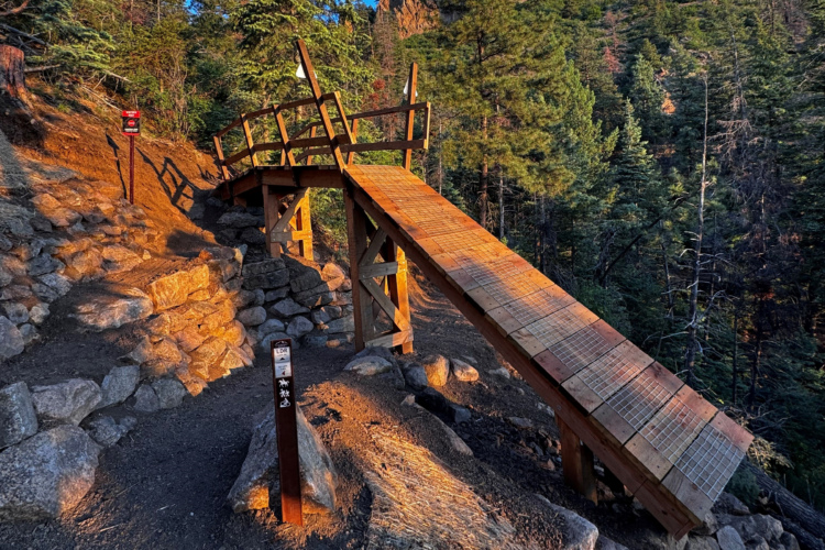 A wooden bridge spans a rocky path in a forested area, surrounded by tall pine trees. The bridge features a textured surface and railings, casting shadows on the ground as the warm light of sunset illuminates the scene. A signpost nearby provides information for hikers, enhancing the natural landscape's accessibility.