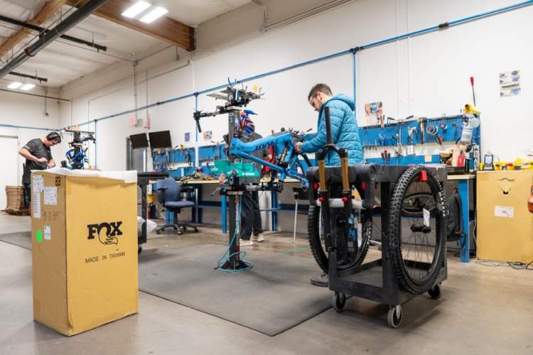 A workshop with two individuals working on bicycles. One person is assembling a bike frame on a repair stand, while the other is unpacking a box labeled "FOX" in the background. The workshop features various tools on a blue workbench and has a clean, organized appearance with concrete flooring and overhead lighting.