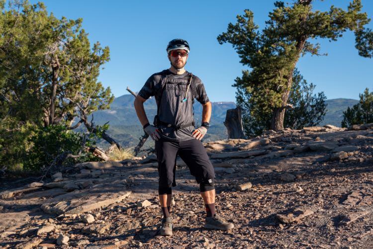 A person wearing a helmet and sunglasses, standing confidently on rocky terrain surrounded by trees and mountains. They are dressed in a short-sleeve athletic shirt, knee-length shorts, and hiking shoes, ready for an outdoor adventure.