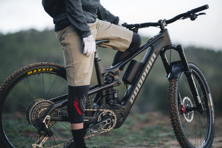 A mountain biker wearing gloves and knee guards stands beside a black electric mountain bike on a dirt trail, surrounded by greenery. The bike features prominent branding and is equipped with a sturdy frame and a multi-gear setup. The rider is dressed in shorts and tall socks, ready for an outdoor adventure.