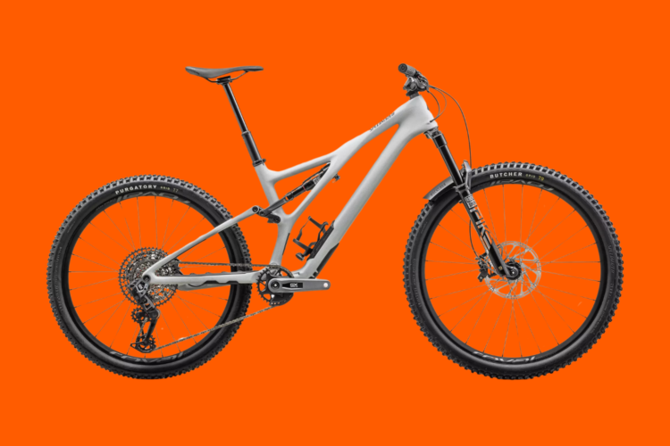 A modern mountain bike with a sleek gray frame, featuring wide tires and suspension components, displayed against a bright orange background.