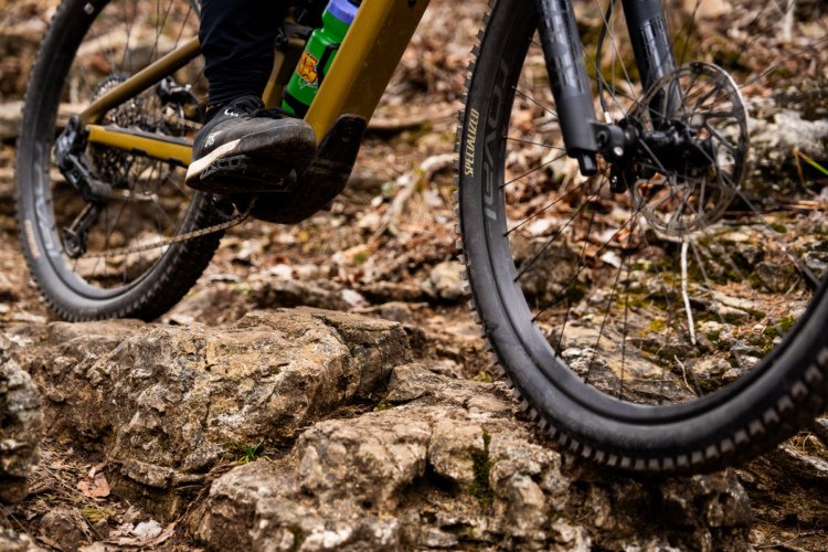 A close-up of a mountain bike navigating rocky terrain, showcasing the bike's front wheel and the rider's foot on the pedal. The background features rough, uneven rocks and patches of brown leaves, indicating a rugged outdoor trail. A water bottle is visible attached to the bike frame.