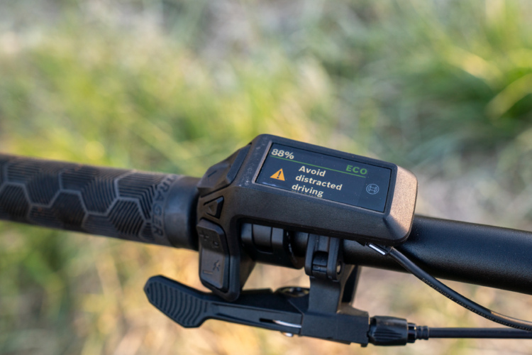 Close-up view of an electric bike handlebar display showing a battery level of 88% and a warning message to "Avoid distracted driving." The handlebar has a textured grip and is set against a blurred green background.