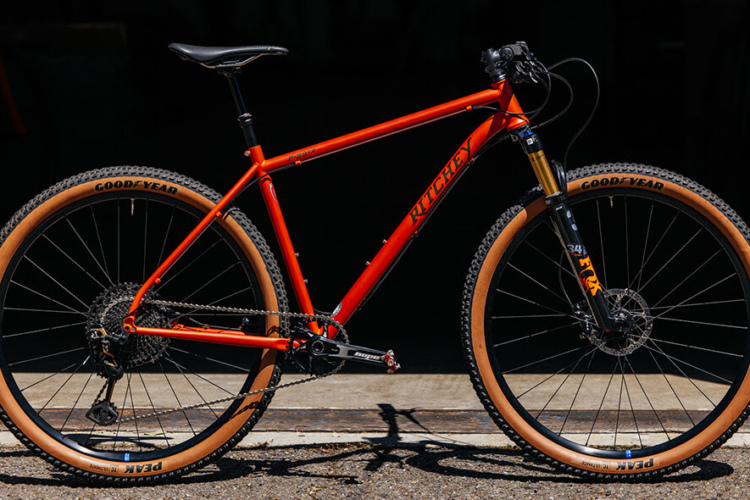 An orange mountain bike with black and brown accents is positioned against a dark background. The bike features a lightweight frame, wide tires with Goodyear branding, and a front suspension fork, indicating its suitability for off-road trails.