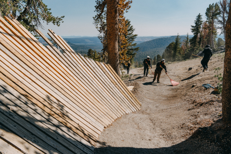 A group of workers are seen maintaining a mountain bike trail, with a wooden structure in the foreground. They are using rakes to smooth out the dirt path, while trees and mountains are visible in the background under a clear blue sky.