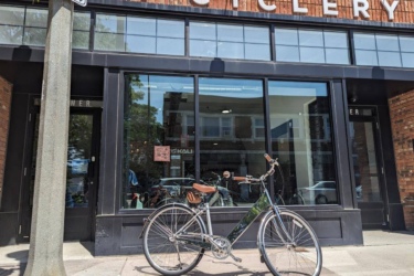 A bicycle is parked outside a brick storefront labeled "City Cyclery." The shop features large windows displaying a bright interior, with a clear blue sky reflected on the glass. A tree branch partially shadows the sidewalk.