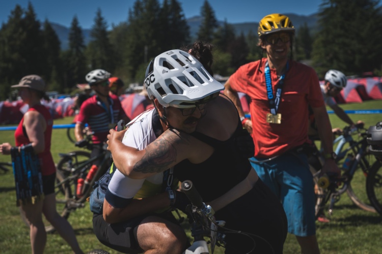 A group of cyclists celebrating in a grassy area, with one person in a white helmet embracing another. The scene features other cyclists in the background, some holding medals and wearing cycling gear. Tents are visible in the distance, and the setting is bright and sunny with trees and mountains in the background.