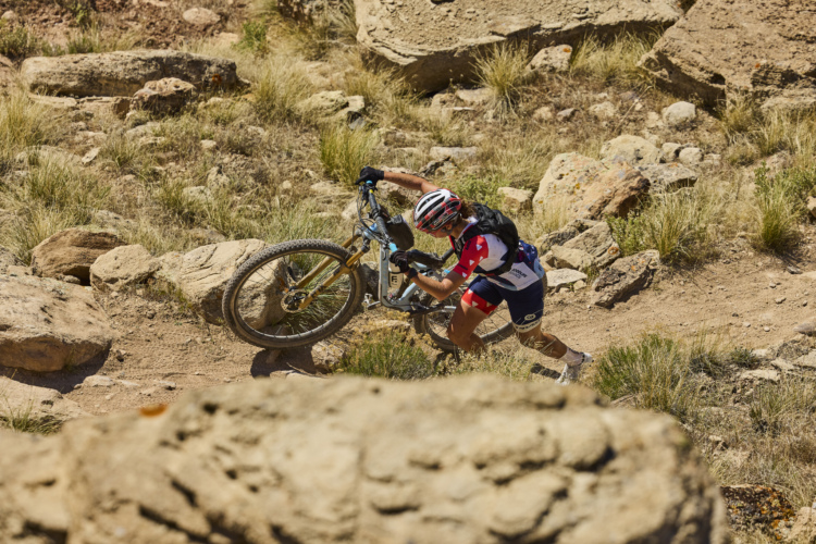 A cyclist navigating a rocky trail, lifting their mountain bike over boulders while surrounded by dry grass and rugged terrain under a clear sky.