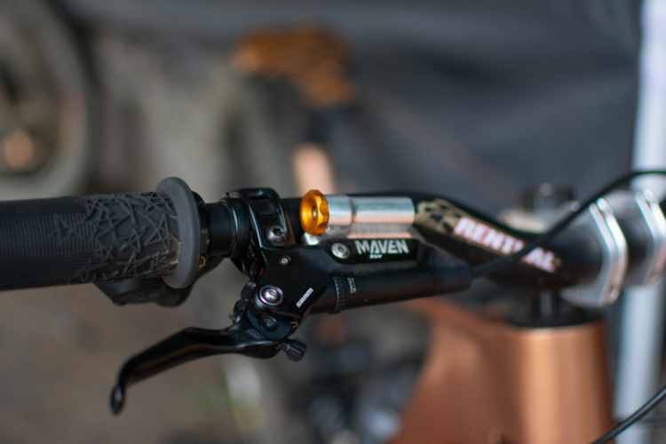 Close-up of a bicycle handlebar showing a textured grip and a brake lever. The handlebars include a MAVEN logo and a shiny metallic component attached, emphasizing the bike's detailed design. Blurred background hints at additional bicycle components.