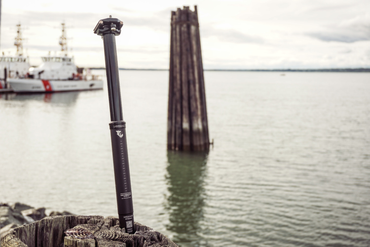 A close-up of a black dropper post resting on a weathered wooden post, with calm water in the background. Two boats are visible in the distance, docked near a series of wooden structures. The sky is overcast, creating a serene atmosphere.