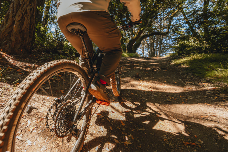 A person riding a mountain bike on a dirt trail surrounded by trees. The photo captures the bike from a low angle, focusing on the rear wheel and the rider's legs. Sunlight filters through the foliage, illuminating the trail ahead.