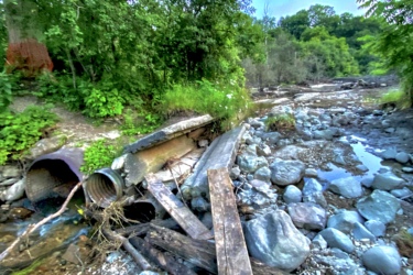 A gravelly streambed surrounded by lush greenery, featuring a broken drainage pipe and wooden planks scattered across the rocky terrain. The sky is bright blue with hints of clouds, suggesting a warm, sunny day. Inverse of kains mountain bike trail.