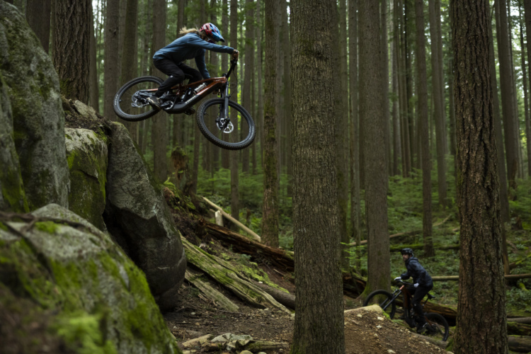 A mountain biker jumps off a rocky ledge in a dense forest, surrounded by tall trees and lush greenery. Another biker is seen in the background, observing the jump.