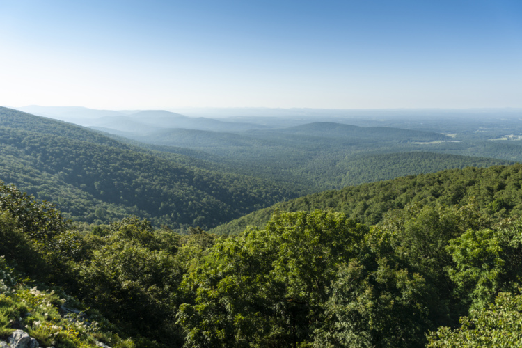 A panoramic view of lush green mountains and valleys under a clear blue sky, showcasing a peaceful natural landscape with rolling hills and dense forestation in the foreground.