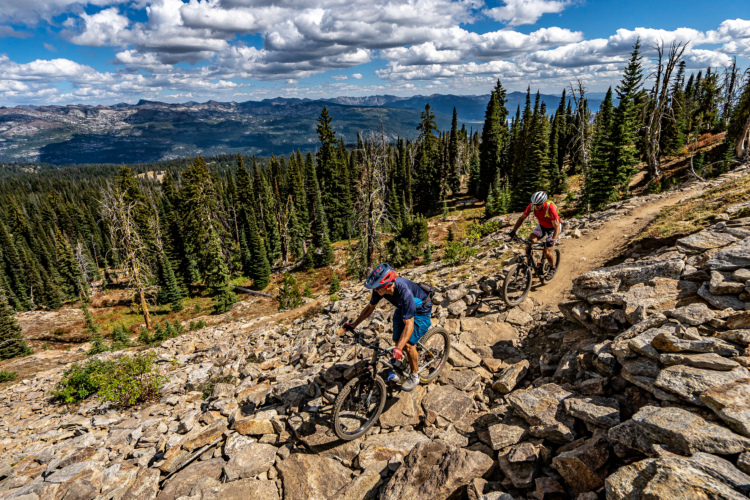 Two mountain bikers navigate a rocky trail in a forested area, surrounded by tall pine trees and scenic mountain views under a partly cloudy sky. The landscape features a mix of rocky terrain and greenery, creating a dynamic outdoor adventure setting.