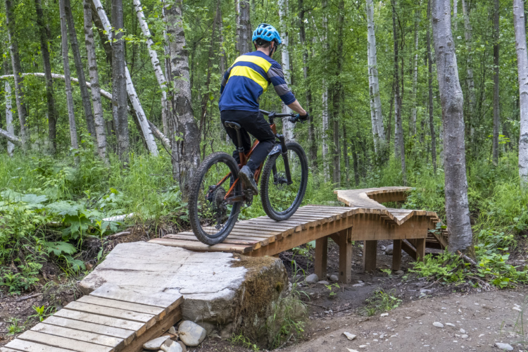 A person riding a mountain bike on a wooden bridge in a lush, green forest. The bridge features a winding design and is surrounded by trees, creating a natural outdoor setting. The cyclist is wearing a blue helmet and a brightly colored jersey, navigating the trail with focus.