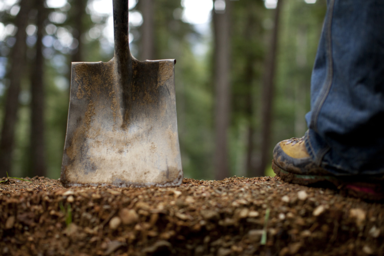 A close-up view of a metal shovel resting on a dirt patch with a person's foot wearing a rugged boot next to it, surrounded by a forest setting in the background.