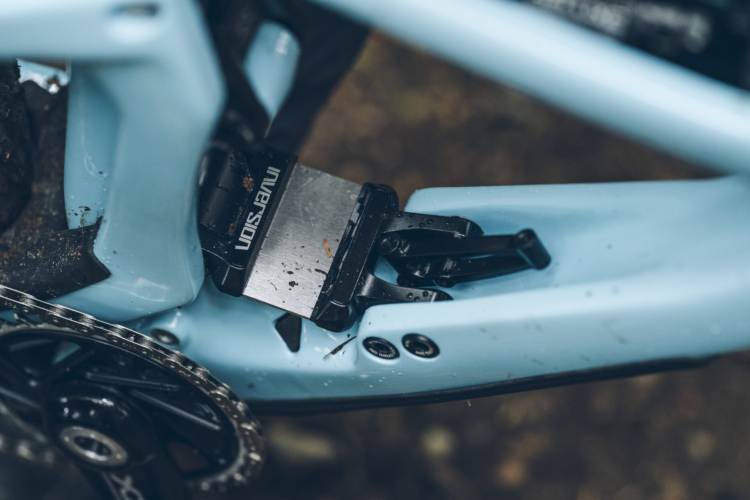 Close-up of a mountain bike's drivetrain, focusing on the derailleur and chainring. The bike features a light blue frame with some dirt smudges, indicating off-road use. The derailleur is partially visible, showcasing mechanical components and the chain.