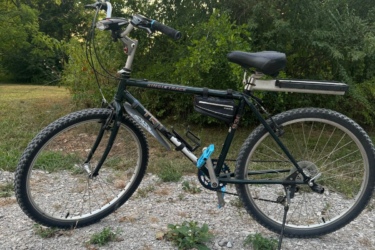 Trek 930: A black mountain bike with blue accents is parked on a gravel path, surrounded by greenery and tall grass. The bike features a singletrack design, with thick tires suited for rough terrain and a comfortable seat. The handlebars are equipped with gear shifters and a small storage bag.