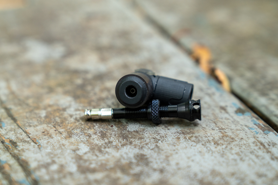 A close-up image of two black mechanical components resting on a weathered, wooden surface, one of which is a small nozzle-like part with a rounded end and the other a cylindrical piece with a metal tip.