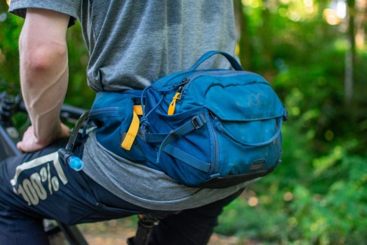 A close-up image of a person wearing a blue hiking or biking waist pack while seated on a mountain bike, with greenery in the background. The pack features various compartments and straps, and the person is dressed in a gray shirt and black pants with a logo.