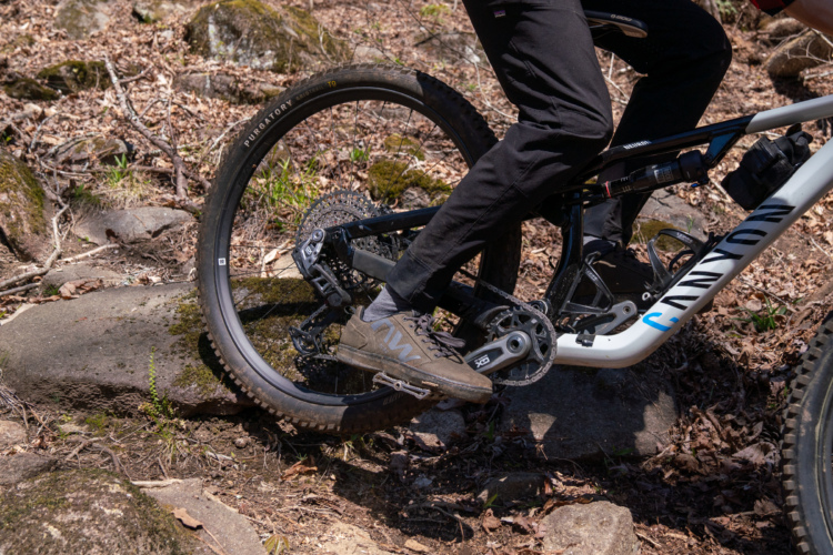 A close-up view of a mountain bike's drivetrain, featuring a rider's foot on the pedal. The bike is navigating over rocky terrain, surrounded by leaves and underbrush. The bike frame is prominently branded with "CANYON," and the tire has a tread pattern suitable for off-road biking.