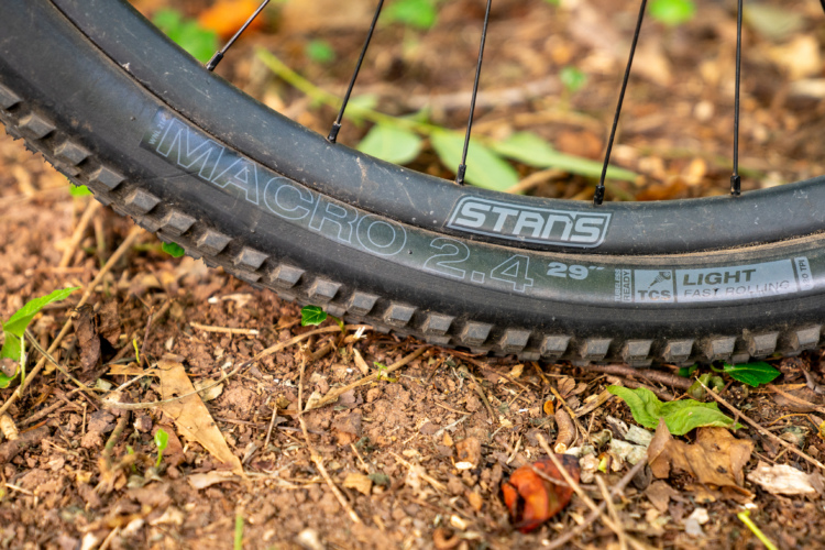 Close-up view of a mountain bike tire resting on dirt and leaves, featuring visible brand markings "Stans" and "Macro 2.4 29." The tire's tread pattern is highlighted, showcasing its design for off-road cycling.