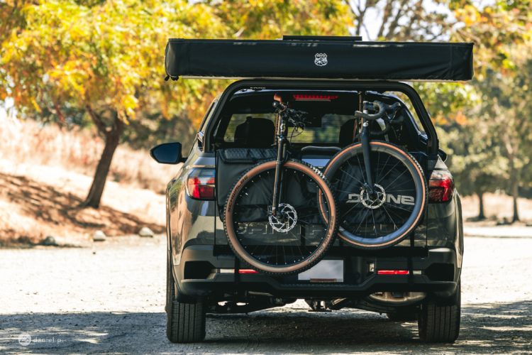 A black SUV parked on a gravel surface with a rooftop tent extended. Two mountain bikes are securely mounted on the back of the vehicle, surrounded by a natural landscape featuring trees with autumn foliage.