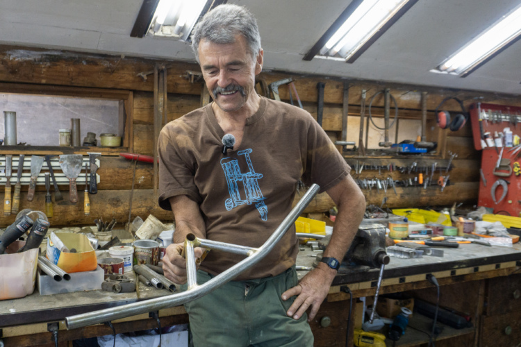 A man stands at a workbench in a workshop filled with tools and materials. He is smiling and holding a bicycle frame made of metal tubing, examining it closely. The workshop has wooden walls and is well-lit, with various tools and containers visible in the background. The man is wearing a brown t-shirt with a graphic design and green pants.