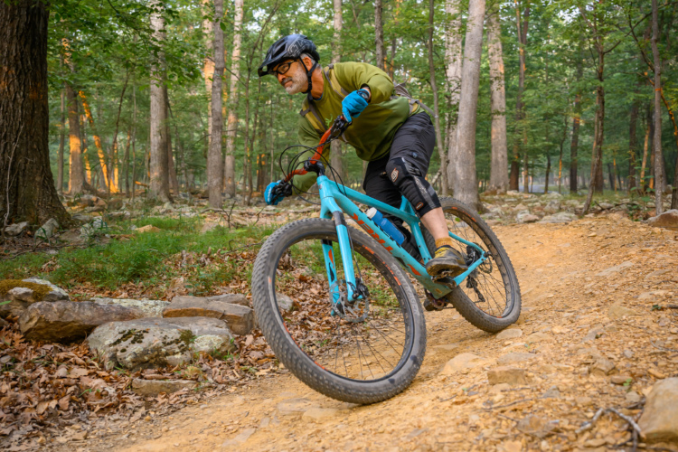A cyclist in a green long-sleeve shirt and black shorts navigates a rocky trail on a blue mountain bike in a wooded area. The scene features tall trees, green foliage, and a dirt path, showcasing an outdoor biking adventure.