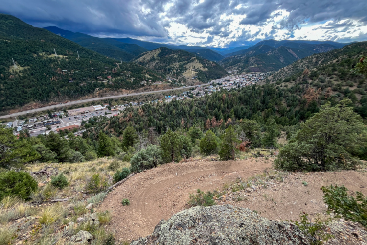A panoramic view of a mountainous landscape with rolling hills and lush greenery, overlooking a small town nestled in the valley below. The scene features a winding dirt path in the foreground, leading down the slope, with the town's buildings visible alongside a road. The sky is overcast with dramatic clouds, adding depth to the picturesque scenery.
