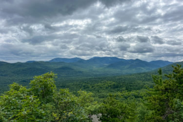 A panoramic view of lush green mountains under a cloudy sky. The landscape features rolling hills covered in dense foliage, with a range of mountains visible in the distance. The clouds are varied in texture, creating a dramatic atmosphere above the vibrant greenery. Blueberry Hill mountain bike trail.