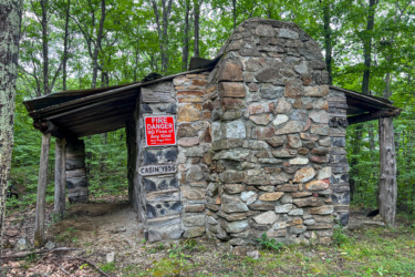 A rustic stone cabin partially nestled in a wooded area, featuring a roof supported by wooden beams. A prominent red sign on the cabin warns of fire danger and prohibits making fires of any kind. The cabin is marked with the year "1996" on the side. Surrounding foliage includes green trees and underbrush, creating a serene natural setting.