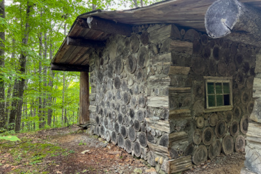 A rustic log cabin partially nestled among dense green trees, featuring a fa&ccedil;ade made of stacked tree rounds and a small window. The surrounding area is covered with leaves and earthy ground, capturing a serene woodland atmosphere. Blueberry Hill mountain bike trail.