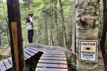 A mountain biker stands thoughtfully on a large rock in a forested area, wearing a helmet and casual riding gear. In the foreground, a wooden ramp curves around a tree, leading to a steep drop. A caution sign reads "CAUTION ADVANCED FEATURE - Ride at your own risk," warning of the challenging terrain ahead. The setting features lush green trees, creating a natural backdrop.