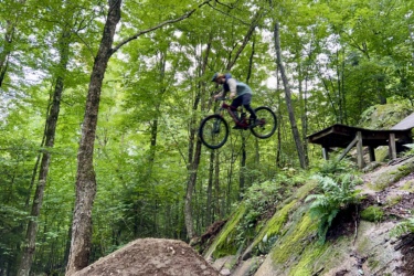 A mountain biker airborne mid-jump over a dirt ramp, surrounded by lush green trees and foliage, in a forested area. Craig Wood Trails mountain bike trail.