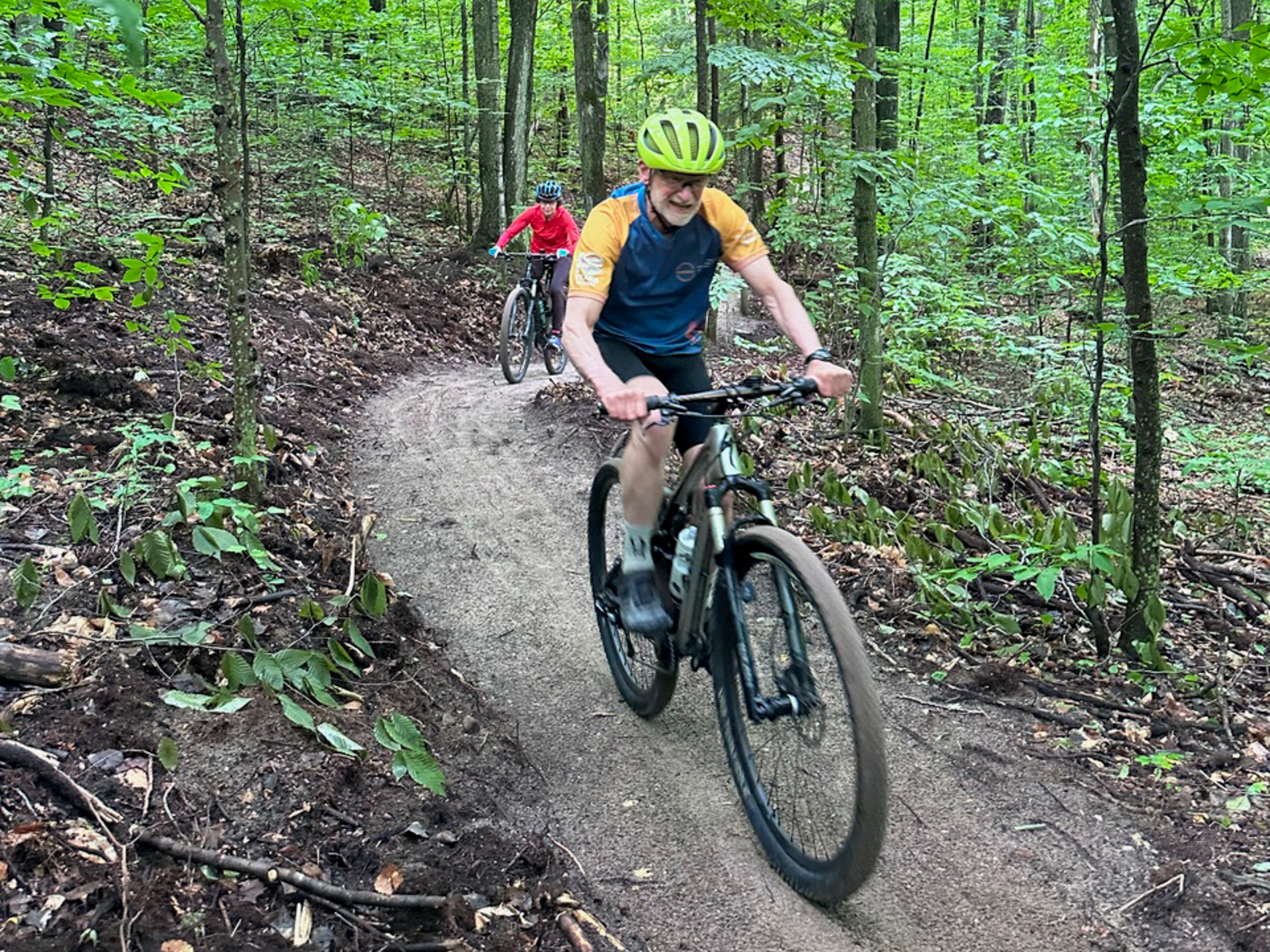 A man in a yellow helmet and colorful cycling shirt rides a mountain bike along a winding trail in a lush green forest. Behind him, another cyclist in a red jacket follows the same path. The scene showcases a vibrant, natural environment with trees and foliage surrounding the trail. Offield Viewlands Mountain Bike Trail mountain bike trail.