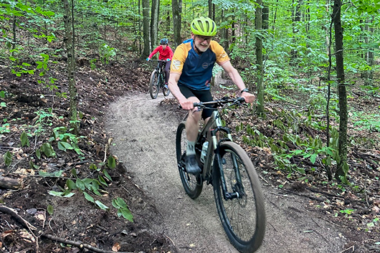 Two cyclists ride on a dirt path through a lush, green forest. One cyclist, wearing a yellow helmet and a blue shirt, leads the way, while the second cyclist, dressed in red, follows closely behind. Bright green leaves and trees surround the trail, creating a vibrant outdoor scene.