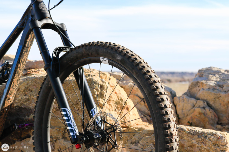Close-up of a mountain bike's front wheel and suspension fork, resting on rocky terrain with a clear blue sky in the background. The tire features deep treads suitable for off-road conditions, and the bike is partially obscured by rocks.